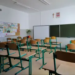 Pusta sala lekcyjna z rzędami krzeseł i biurek, tablicą i dziecięcymi rysunkami na ścianie.