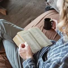 Osoba z kubkiem w dłoni czyta otwartą książkę, siedząc na kanapie.