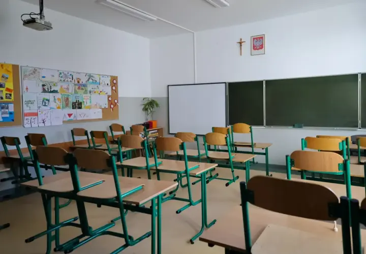 Pusta sala lekcyjna z rzędami krzeseł i biurek, tablicą i dziecięcymi rysunkami na ścianie.