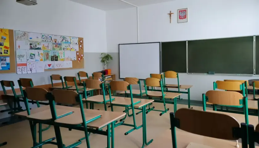 Pusta sala lekcyjna z rzędami krzeseł i biurek, tablicą i dziecięcymi rysunkami na ścianie.