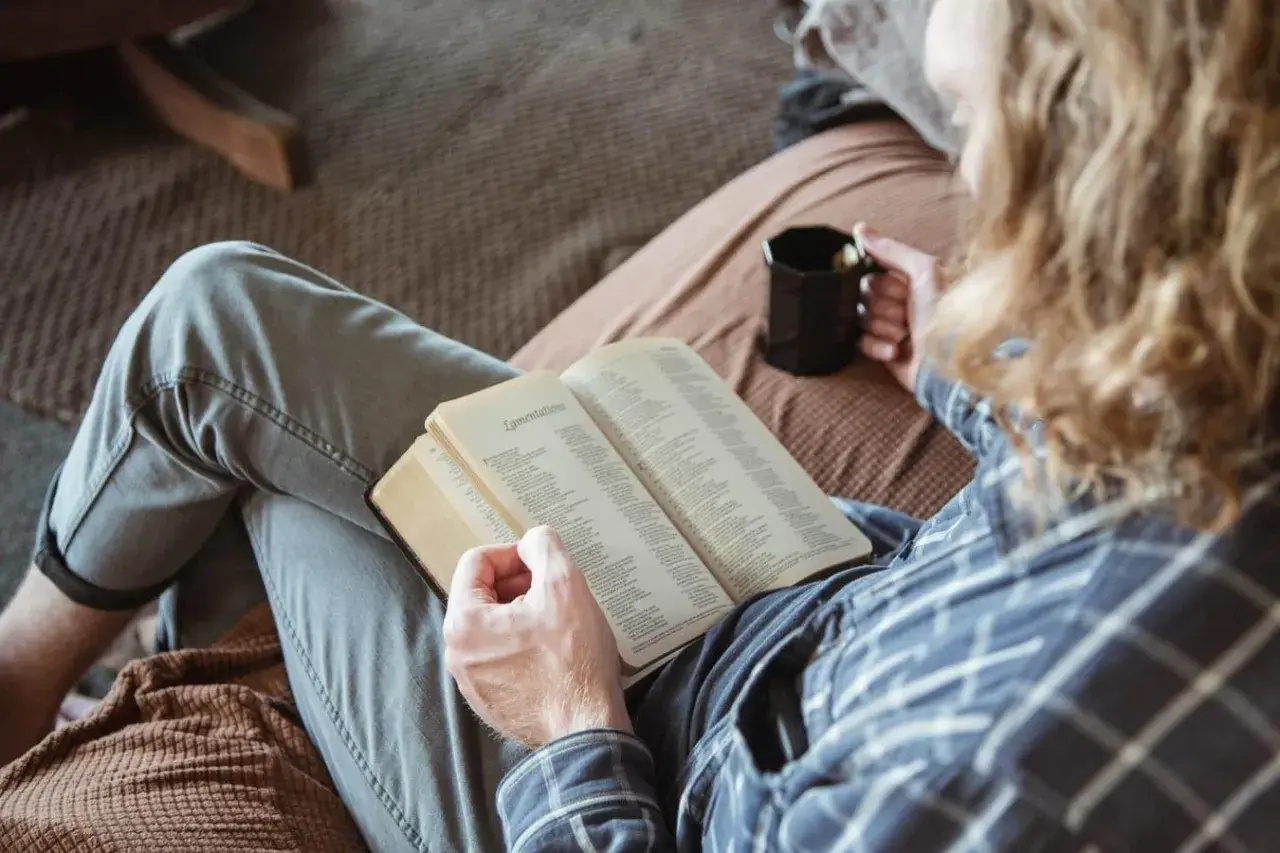 Osoba z kubkiem w dłoni czyta otwartą książkę, siedząc na kanapie.