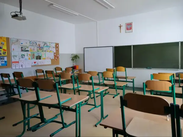 Pusta sala lekcyjna z rzędami krzeseł i biurek, tablicą i dziecięcymi rysunkami na ścianie.